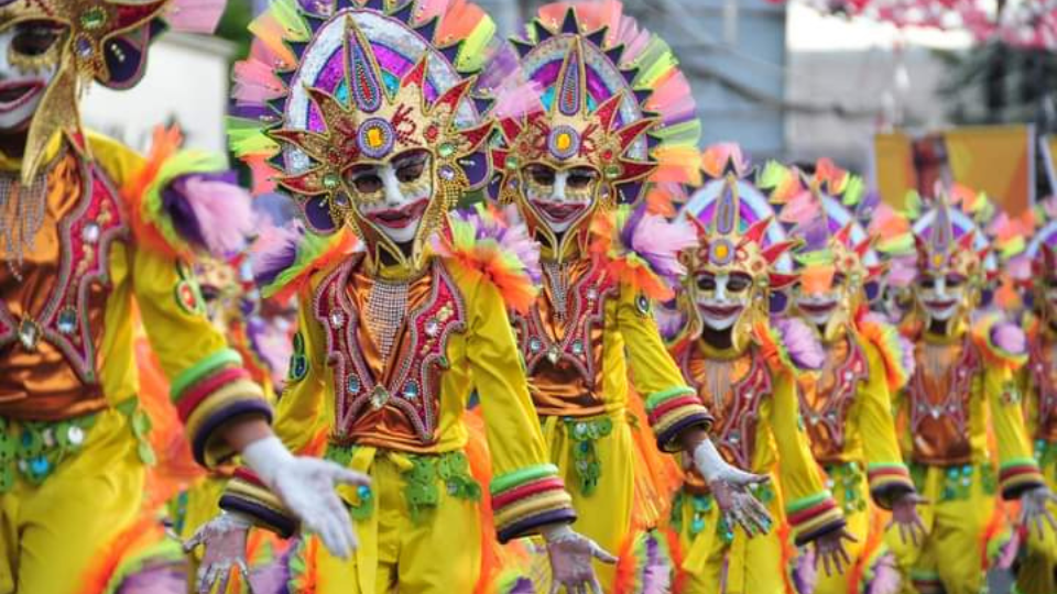 MassKara Festival Article Feature Image. A parade of festival dancers wearing white masks with gold brocade under the eyes and a red painted smiling mouth. A gold crown head piece with red fire tips and yellow square at the center. costumes of yellow material with red, gold, green and black brocade cuffs and chest pieces